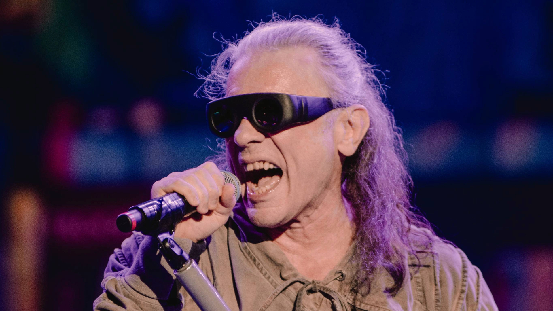 Bruce Dickinson, vocalista do Iron Maiden, cantando ao microfone durante apresentação ao vivo, usando óculos escuros e sob iluminação azul e roxa.