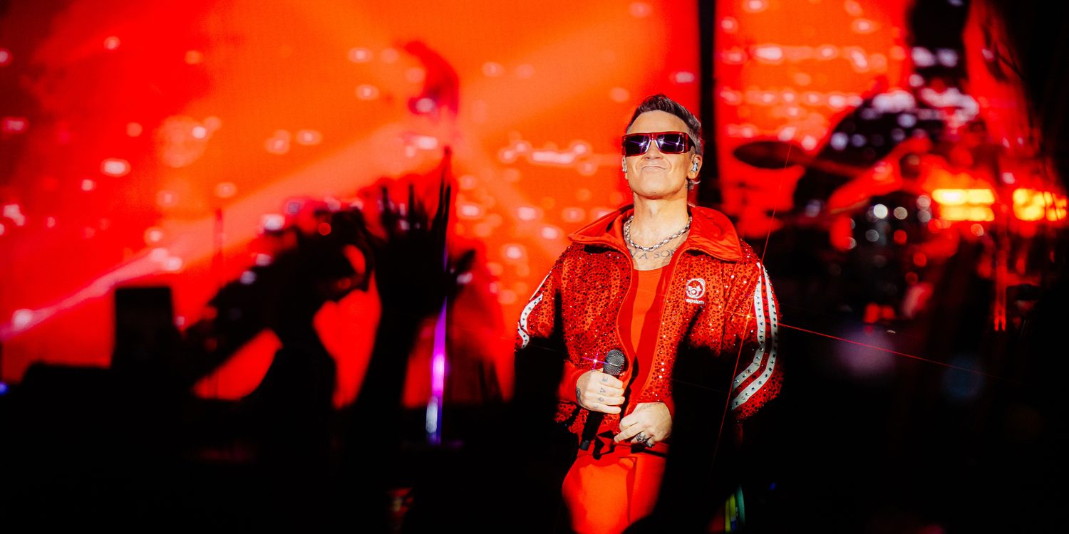 Robbie Williams usa roupa vermelha e óculos escuros enquanto segura um microfone durante show no Nubank Parque, em São Paulo.