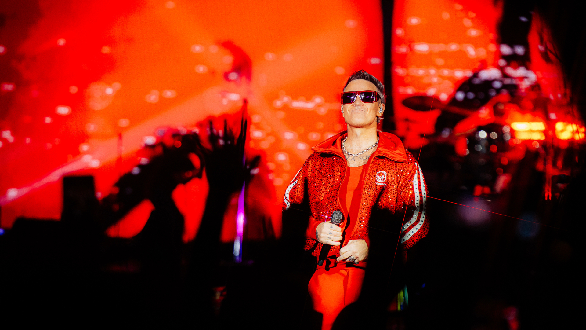 Robbie Williams usa roupa vermelha e óculos escuros enquanto segura um microfone durante show no Nubank Parque, em São Paulo.