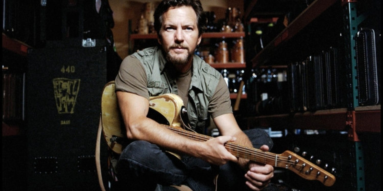 Eddie Vedder sentado com uma guitarra elétrica no colo, em um ambiente com equipamentos musicais e estantes ao fundo.