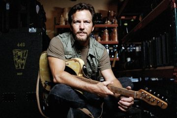 Eddie Vedder sentado com uma guitarra elétrica no colo, em um ambiente com equipamentos musicais e estantes ao fundo.