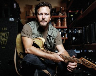 Eddie Vedder sentado com uma guitarra elétrica no colo, em um ambiente com equipamentos musicais e estantes ao fundo.