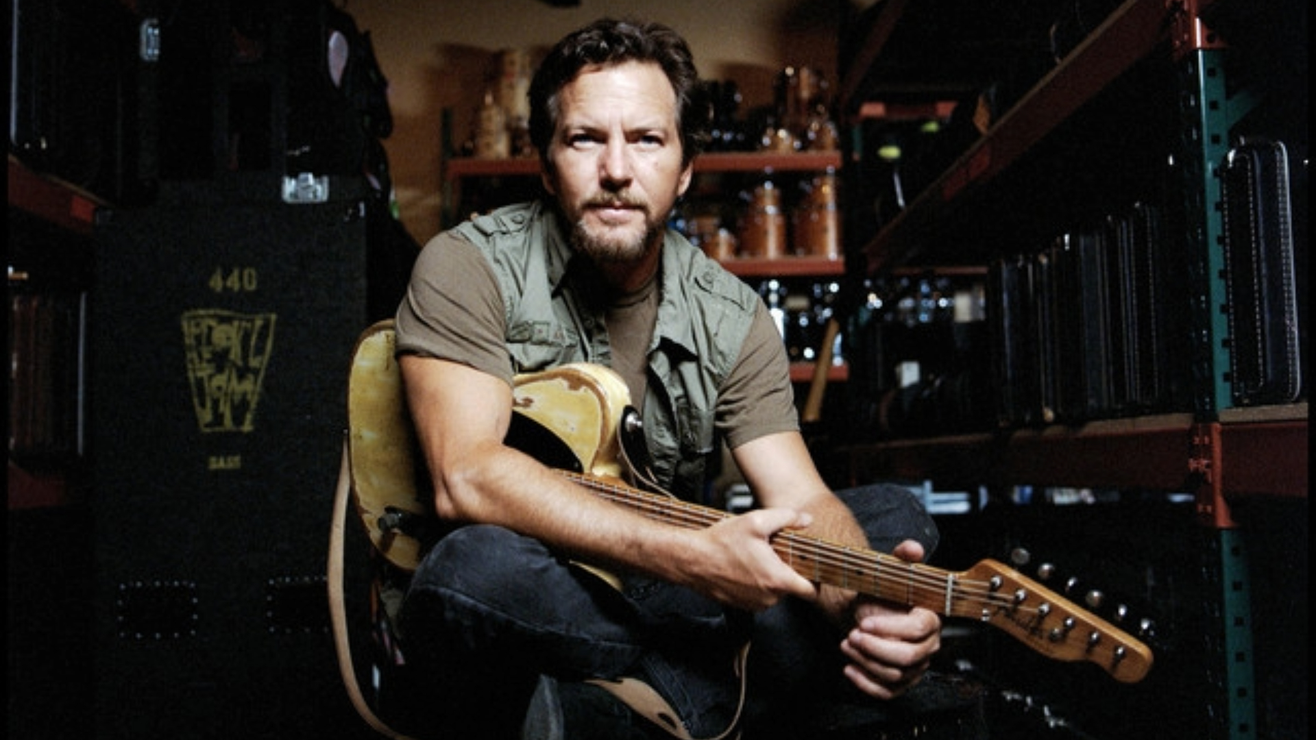 Eddie Vedder sentado com uma guitarra elétrica no colo, em um ambiente com equipamentos musicais e estantes ao fundo.