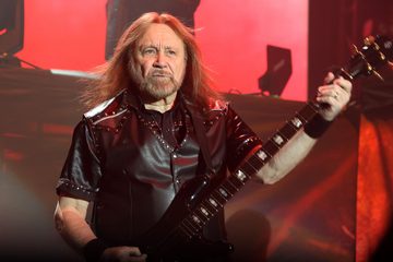 Ian Hill, baixista do Judas Priest, tocando baixo durante apresentação ao vivo, com iluminação vermelha ao fundo.
