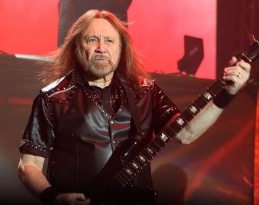 Ian Hill, baixista do Judas Priest, tocando baixo durante apresentação ao vivo, com iluminação vermelha ao fundo.