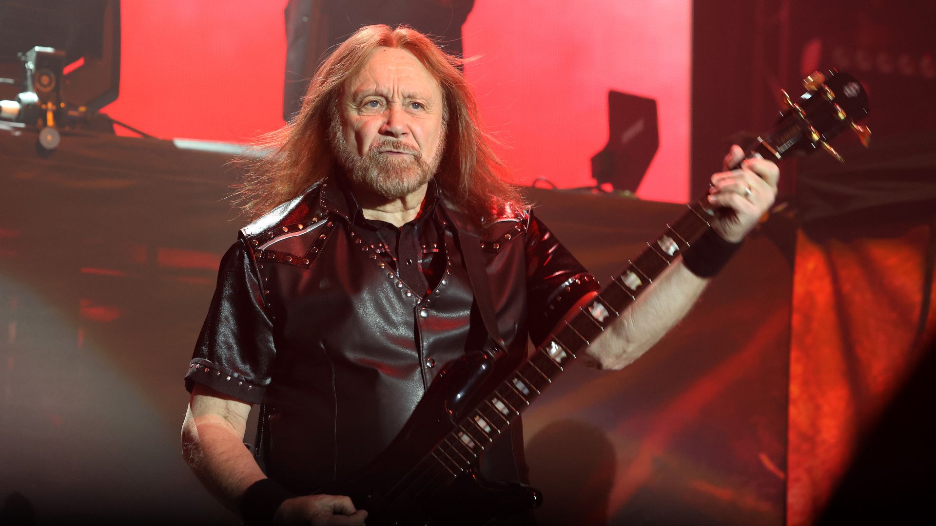 Ian Hill, baixista do Judas Priest, tocando baixo durante apresentação ao vivo, com iluminação vermelha ao fundo.