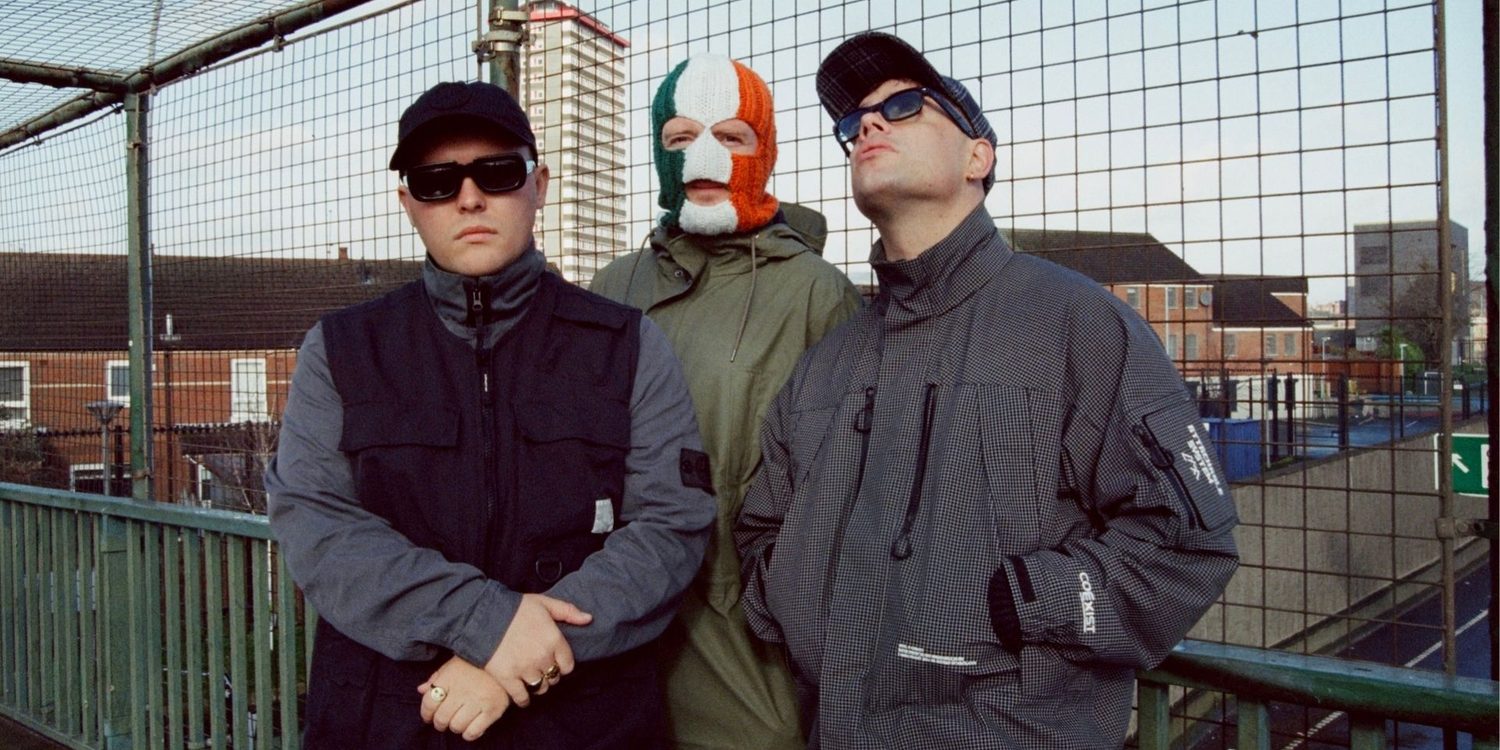 Integrantes do KNEECAP, trio de hip-hop de Belfast, posam em uma passarela cercada por grades, com um dos músicos usando uma balaclava nas cores da bandeira irlandesa.