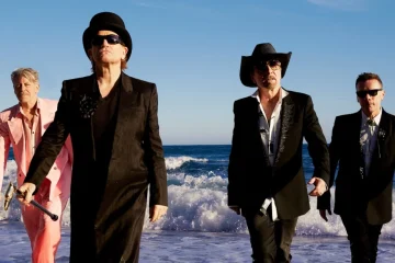 Integrantes do U2 caminham pela praia usando ternos, chapéus e óculos escuros em fotografia de Viviane Sassen.