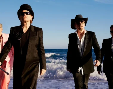 Integrantes do U2 caminham pela praia usando ternos, chapéus e óculos escuros em fotografia de Viviane Sassen.