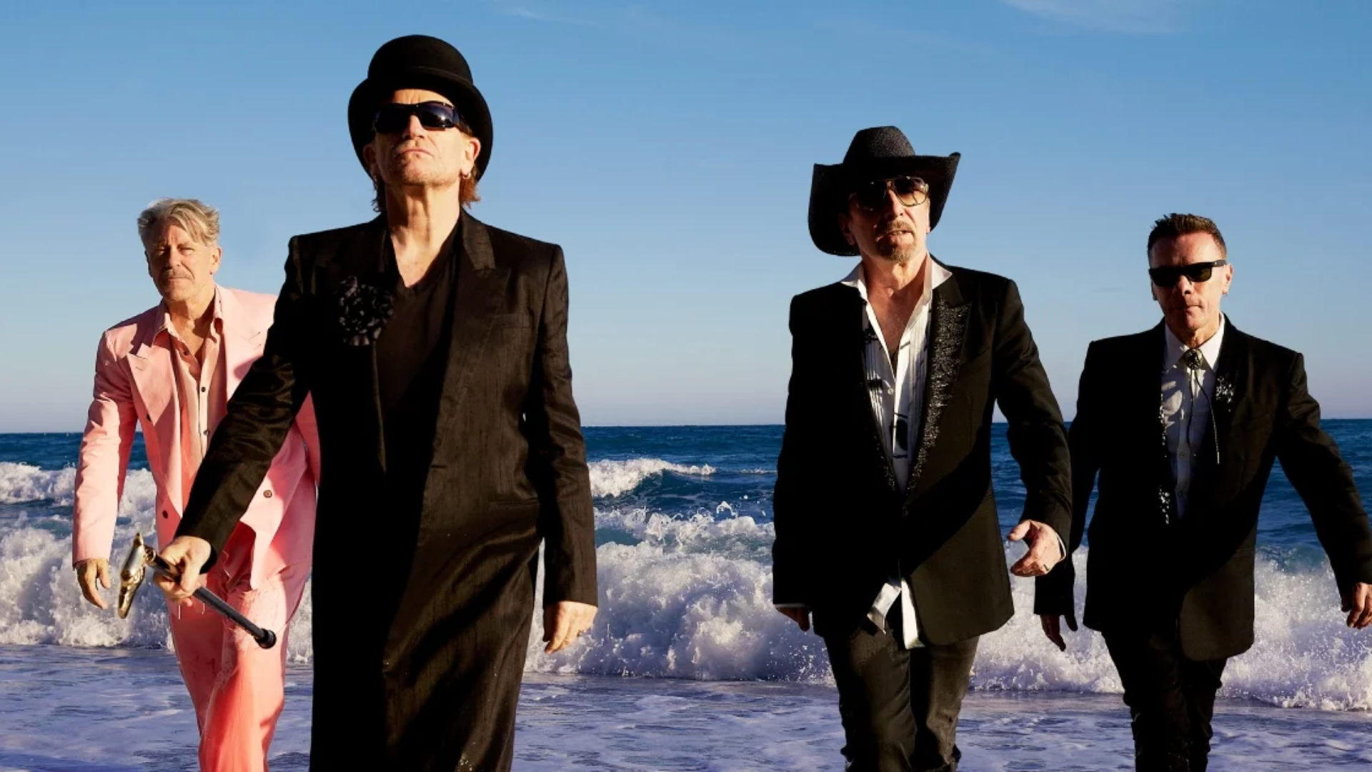 Integrantes do U2 caminham pela praia usando ternos, chapéus e óculos escuros em fotografia de Viviane Sassen.